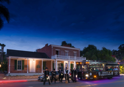 Ghosts & Gravestones tour guides standing in front of a Ghosts & Gravestones trolley filled with guests in front of the Whaley House, a brick historic looking house made of brick with windows and shutters and a porch on the second floor