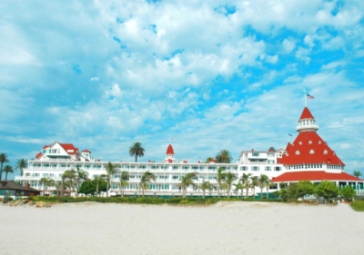 A beachside wide-angle view of the Hotel del Coronado with sand in the foreground and an arrangement of palm trees