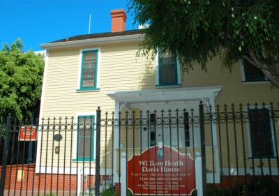 The exterior of the historic Davis-Horton House in San Diego behind a tall iron fence painted a very pale green or yellow constructed in the saltbox style of the mid 1800s