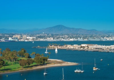 A very wide view of the Ferry Landing in San Diego where you can see a public park with a beach, a naval base, the San Diego bay with several sailboats floating around and mountains in the distance