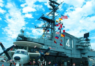 A twin engine navy prop plane parked next to the flag bridge, adorned with different colorful flags and the primary flight control area which has several observation decks in San Diego