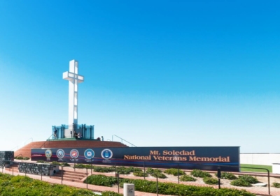 San Diego's Mount Soledad National Veterans Memorial with a long wall where all 5 logos of the military branches are displayed along with a tall white crucifix mounted at the site's highest point