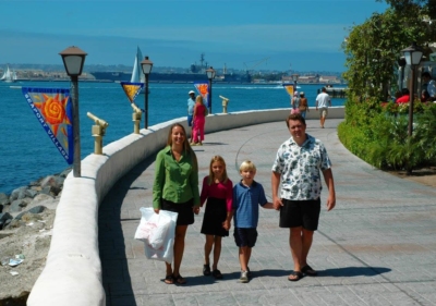 An observation area in San Diego's Seaport Village showing a family of four smiling, telescopes for public use and a large naval vessel in the distance